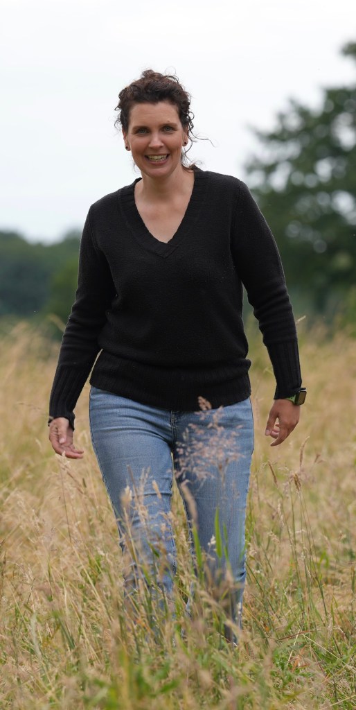 Eine Frau mit lockigem Haar trägt einen schwarzen Pullover und Jeans, während sie durch ein Feld mit hohen Gräsern geht und lächelt.